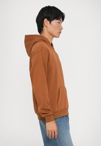 Rjava kapuzen sweatshirt s sprednjim žepom, mehkega teksture in rebrastimi manšetami. Nosimo ga z svetlo modrimi kavbojkami.