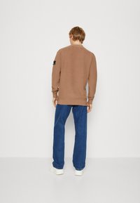 Brązowy sweter z dzianiny z długimi rękawami i średnim okrągłym dekoltem, zestawiony z niebieskimi dżinsami i białymi sneakersami. Zawiera detal z logo.