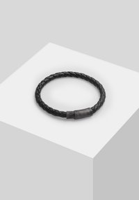 Mustmust ristmik mustast nahast, millel on tekstureeritud metallist kinnitus. Ümmargune kuju, sile pind ja minimalistlik disain.