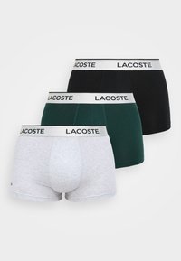 Tre par med Lacoste-boxershorts i grå, mørkegrønn og svart, med en hvit midjebånd med gjentatt "LACOSTE" merke. Myk bomullsstoff.