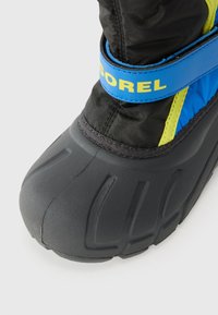 Fekete szigetelt bakancs, texturált gumitalppal, kék állítható pánttal, rajta "SOREL" felirattal, és kétszínű dizájnnal kék és sárga színekben.