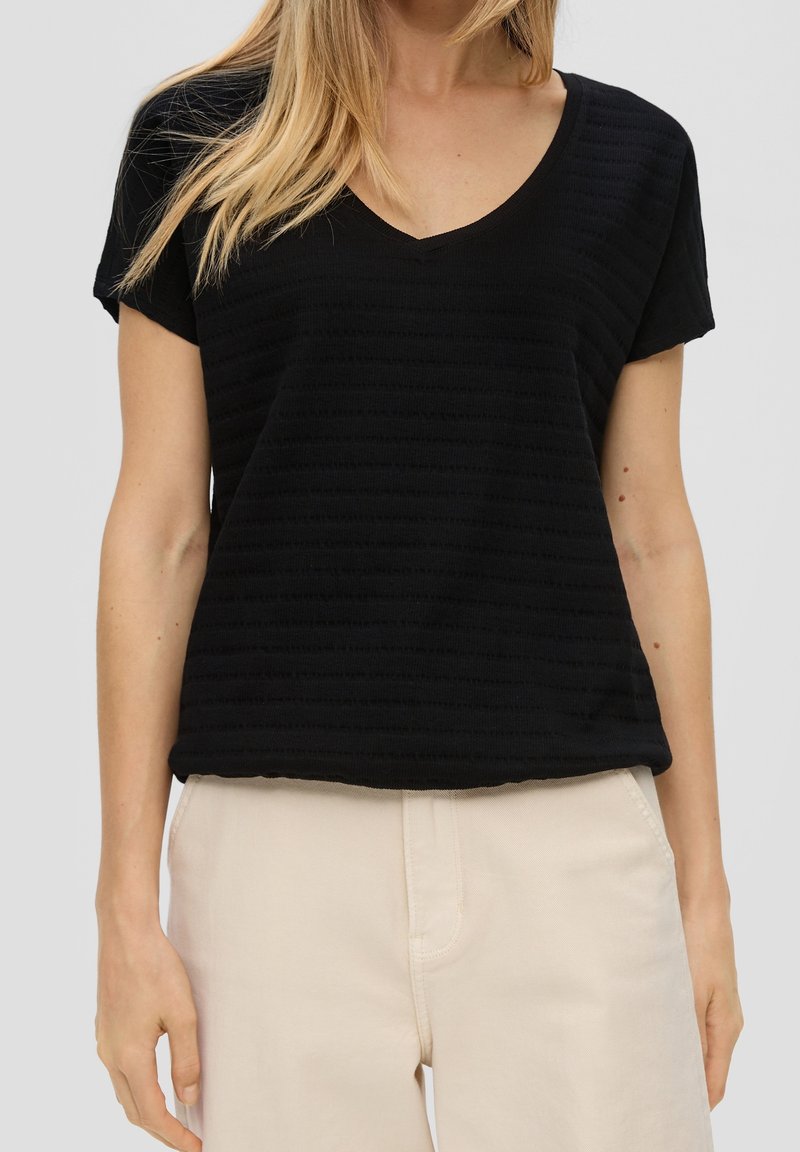 Femme portant un t-shirt noir texturé à manches courtes et col en V, et un pantalon beige clair, debout avec les bras détendus le long du corps.