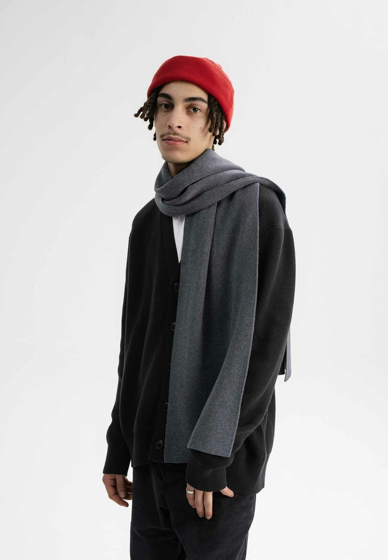 Zwarte cardigan met knopen, grijze sjaal om de nek, rode gebreide muts, witte shirt, donkere broek, gladde textuur, casual outfit.