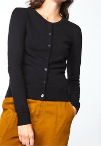 Kvinna iförd en åtsittande svart knappad cardigan och senapsgula byxor, med en hand i fickan mot en vit bakgrund.