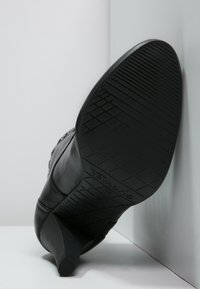 Zapato de tacón negro con suela de goma texturizada. Presenta un material superior liso y un tacón afinado. Marca visible en el lateral de la suela.