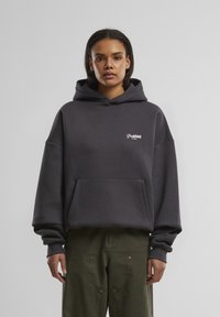 Mørkegrå oversized hoodie lavet af blødt stof, med en frontlomme og et lille hvidt logo på brystet, samt hætte.