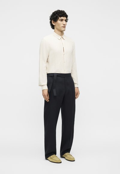 Νέος άνδρας στέκεται, φοράει κρεμ πουκάμισο με μακριά μανίκια και κουμπιά, μαύρο ψηλόμεσο παντελόνι με δεσίματα στο πλάι και μπεζ μοκασίνια.