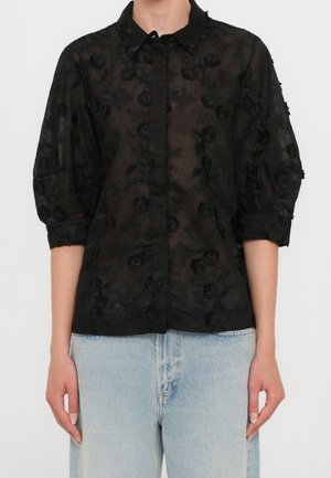 Kvinde iført en gennemsigtig sort bluse med blomsterbroderi og trekvartlange pufærmer, kombineret med lyseblå jeans med højtaljet pasform.
