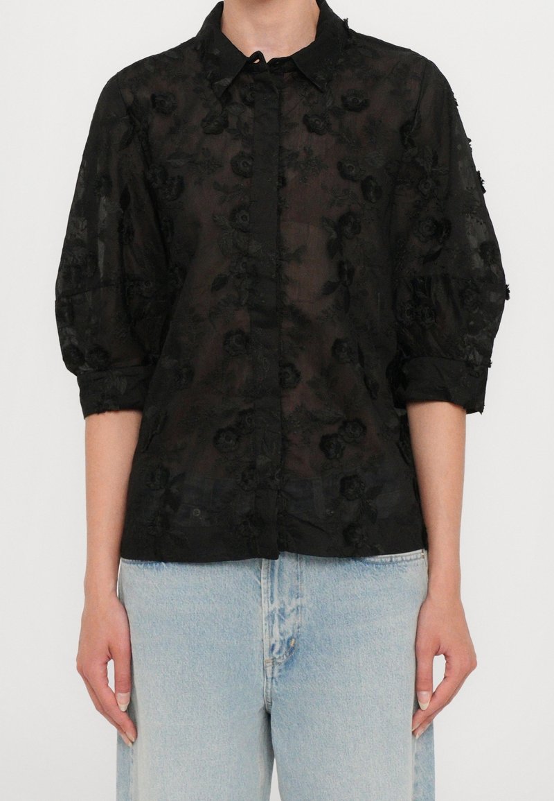Femme portant un chemisier noir transparent avec broderie florale et manches bouffantes trois quarts, associé à un jean taille haute bleu clair.