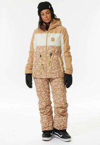 Giacca e pantaloni da snowboard in beige e crema con motivi geometrici. La giacca ha tasche con zip e cordini regolabili. Beanie nero e guanti.