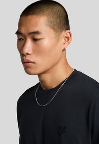 Camiseta negra de manga larga con cuello redondo que presenta un pequeño logo bordado. Un delgado collar de cadena plateada adorna el cuello.