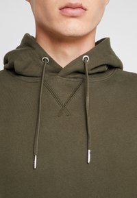Olijfgroene hoodie gemaakt van zacht stof met een gestructureerde afwerking. Heeft een trekkoord met zilverkleurige metalen uiteinden en een gestikte X-patroon op de borst.