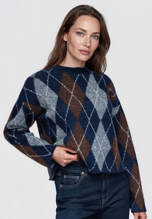 Une femme porte un pull à motif argyle bleu marine, gris et marron, avec un jean foncé, sur un fond clair uni.