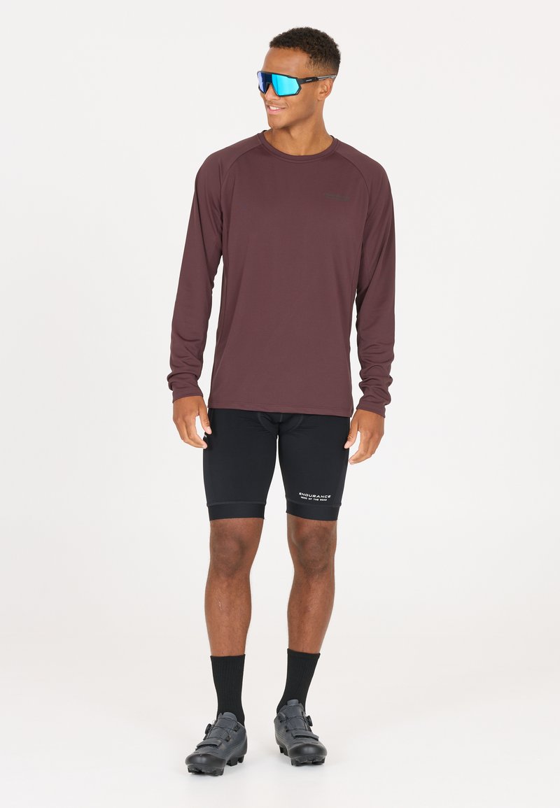 Langärmliges, kastanienfarbenes Sport-Shirt, schwarze Radshorts, schwarze Schuhe und blaue, reflektierende Sonnenbrille; glattes Material, figurnahes Design und Logodetail.