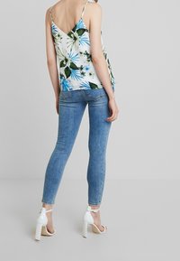 Blomstrig cami-top med gröna och blå mönster, V-back design, i kombination med ljusblå snäva jeans och vita högklackade sandaler.