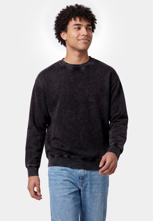 Junger Mann mit lockigem Haar, der einen schwarzen Sweatshirt und hellblaue Jeans trägt, steht und schaut leicht zur Seite mit einem subtilen Lächeln.