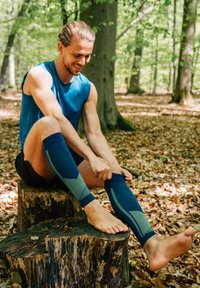 Manguitos de compresión en azul marino, con un patrón geométrico texturizado y acentos acanalados, usados por una persona sentada sobre un tronco en el bosque.