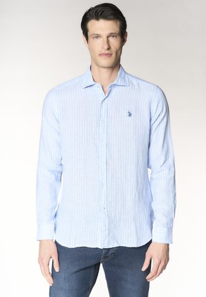 Uomo che indossa una camicia azzurro chiaro a righe verticali e jeans scuri, in piedi davanti a uno sfondo chiaro uniforme.