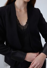 Blazer nero con una leggera lucentezza, caratterizzato da polsini simili alla pelle e una vestibilità su misura. Sotto, una maglia nera con rifiniture in pizzo.