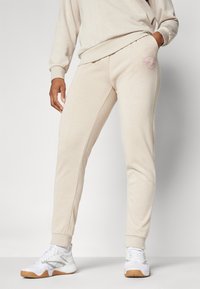 Beige trenirke s ožjim krojem in rebrastimi manšetami. Imajo logo v roza barvi na levem boku. Narejene so iz mehkega, teksturiranega materiala.