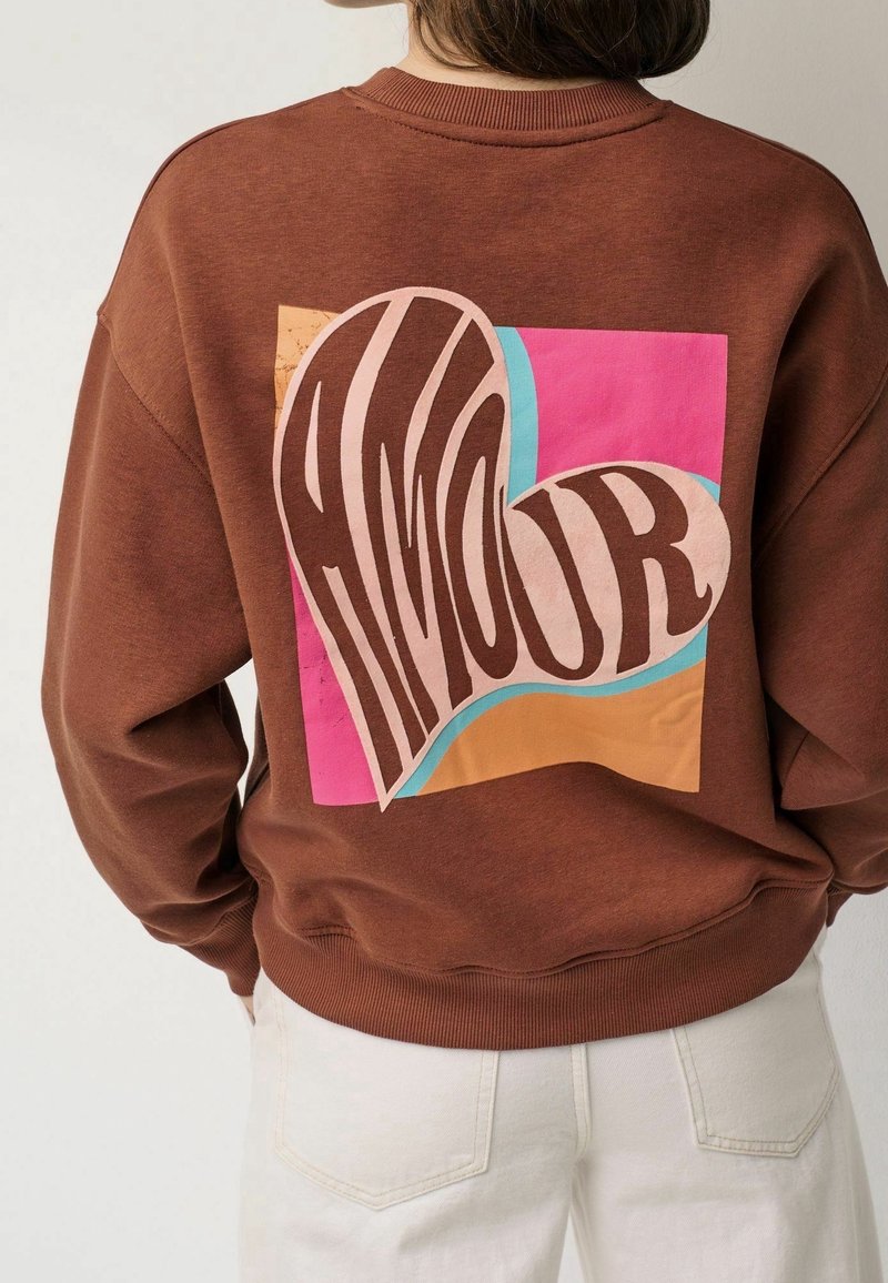 Personne portant un sweat-shirt marron avec un motif abstrait coloré en forme de cœur et le mot « AMOUR » dans le dos, assorti à un pantalon clair.