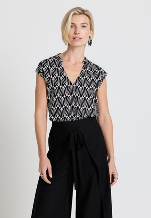 Donna bionda che indossa un top nero e bianco con scollo a V e maniche corte, abbinato a pantaloni neri a gamba larga e vita alta, legati in vita.