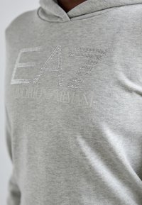 Sivá mikina s kapucňou z textúrovanej tkaniny. Obsahuje nápisy "EA7" a "Emporio Armani" s kryštálovými ozdobami. Jednoduchý dizajn s voľným strihom.