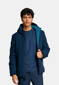 Giacca navy con zip e cappuccio, con fodera a contrasto color teal. Realizzata in materiale sintetico, presenta una vestibilità comoda e polsini elastici.