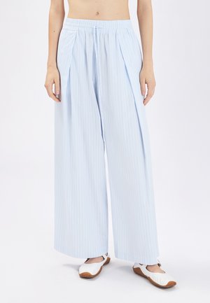 Donna che indossa pantaloni a gamba larga azzurri a righe sottili con vita elastica e scarpe bianche basse su sfondo bianco.