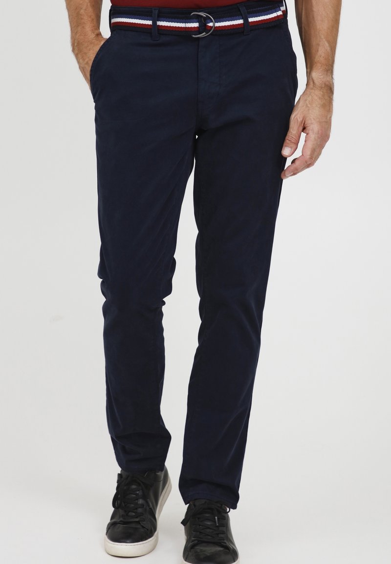 Homme portant un pantalon bleu marine slim avec une ceinture rayée et des baskets noires, debout les mains dans les poches.
