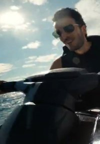 Un homme portant des lunettes de soleil et un gilet de sauvetage fait du jet ski sur une eau ensoleillée avec des nuages dans le ciel.