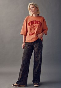 Oranž toonis suur T-särk, millel on punasega kujundus "Windsor Wild Cats"; kombineeritud pruunide leopardimustriliste laiade pükstega ja helepruunide tossudega.