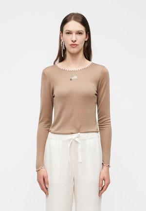 Donna con capelli castani lisci, che indossa una maglia beige a maniche lunghe con rifiniture rosa e pantaloni bianchi con coulisse, in piedi davanti a uno sfondo semplice.
