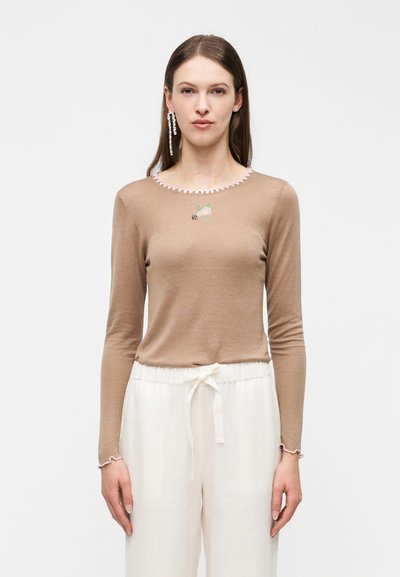 Femme aux cheveux bruns raides portant un haut à manches longues beige avec un bord rose et un pantalon blanc à cordon, debout devant un fond uni.