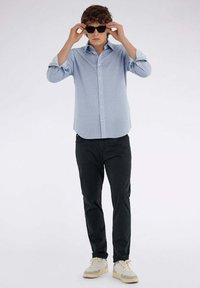 Camicia button-up azzurro chiaro con un motivo testurizzato, abbinata a pantaloni neri e sneakers con accenti beige; occhiali da sole aggiungono un tocco elegante.