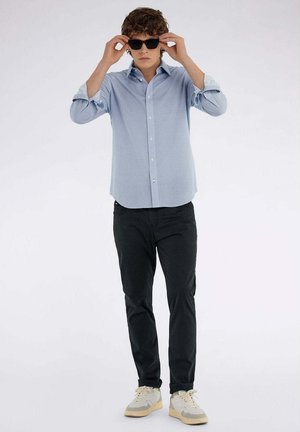 Camicia button-up azzurro chiaro con un motivo testurizzato, abbinata a pantaloni neri e sneakers con accenti beige; occhiali da sole aggiungono un tocco elegante.