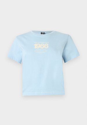 Młoda kobieta o kręconych włosach, ubrana w jasnoniebieski krótki top i jasnoszare dżinsy z wysokim stanem, stoi na jednolitym tle.