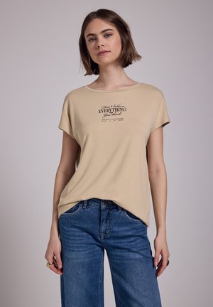 T-shirt beige à manches courtes avec un col rond, présentant un imprimé de texte noir au centre. Associé à un pantalon en jean large bleu. Tissu en coton doux.