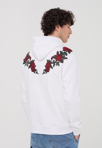 Felpa bianca con ricamo di rose rosse sulle spalle e sul retro, realizzata in tessuto morbido con vestibilità comoda e cappuccio regolabile.