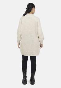 Długi, kremowy, dziergany sweter z wysokim kołnierzem, prążkowaną fakturą i opuszczonymi ramionami. Modelka ma na sobie czarne legginsy i buty.