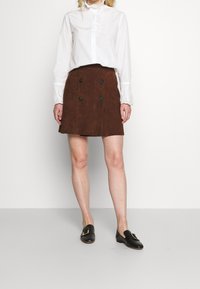 Brauner Wildleder-A-Linien-Rock mit doppelt-knopfeligem Detail, kombiniert mit einer weißen Bluse mit Rüschenkragen und Spitzenbesatz sowie schwarzen Slippern.
