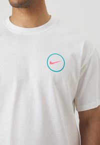Homme portant un t-shirt blanc avec un petit logo Nike swoosh rose à l'intérieur d'un cercle bleu sur le côté gauche de la poitrine.