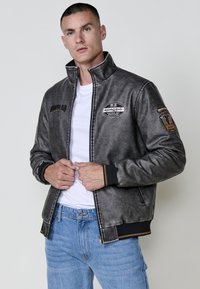 Chaqueta bomber de imitación cuero gris, con un alto collar, parches bordados y puños acanalados. Cierre frontal con cremallera y bolsillos laterales.
