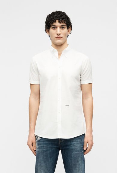 Jeune homme aux cheveux noirs courts et bouclés, portant une chemise blanche à manches courtes avec boutons et un jean délavé bleu, debout devant un fond uni.