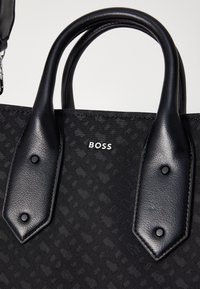 Sac à main noir texturé avec des poignées en cuir noir lisse et un logo argenté "BOSS" centré entre les poignées.