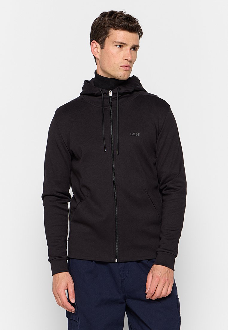 Man in een zwarte zip-up hoodie met het "BOSS" logo op de borst, gecombineerd met marineblauwe cargo pants, die iets opzij kijkt.
