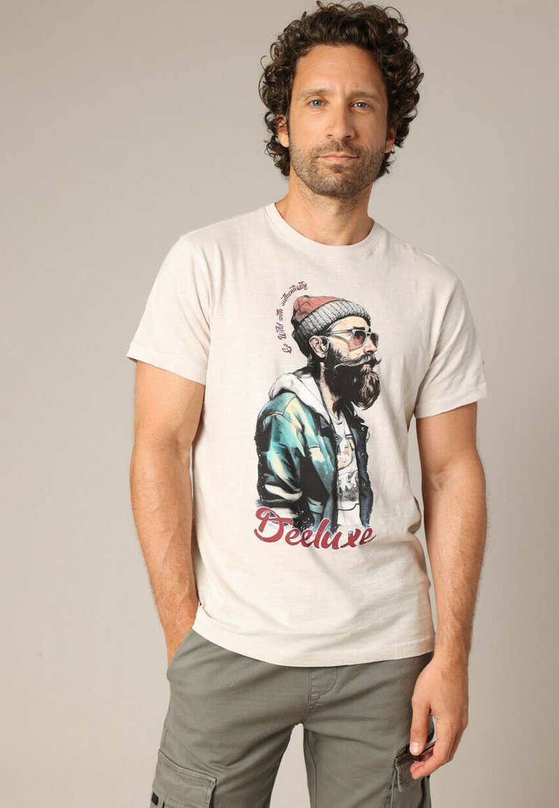 T-shirt à manches courtes couleur crème avec un motif représentant un homme barbu portant des lunettes de soleil et un bonnet rouge, avec le texte "Deluxe" en rouge.