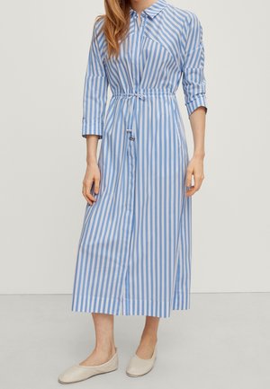 Kvinde iført en midi-kjole med blå og hvide lodrette striber, 3/4-ærmer, krave, knapper foran og snøre i taljen, kombineret med beige flade sko.