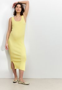 Yellow ribbed midi dress with a scoop neckline and side slit; paired with metallic flat shoes. Simple, form-fitting design.