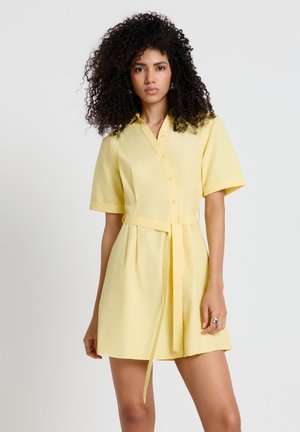 Donna con i capelli ricci che indossa un vestito giallo a maniche corte con bottoni e cintura annodata in vita, in piedi contro uno sfondo chiaro e uniforme.